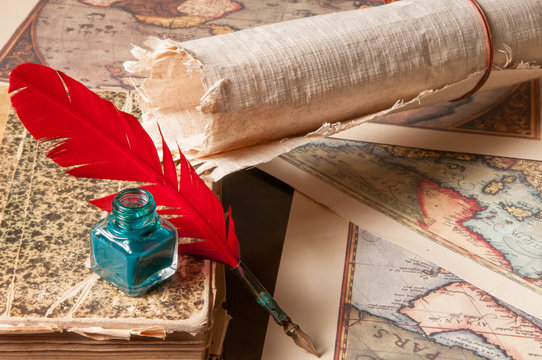 Red quill pen, a green inkwell and a rolled papyrus sheet with some old maps on a wooden table