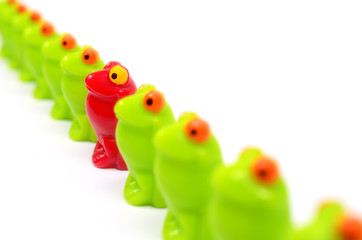 Line of green toy frogs. Among them, one red frog. Close up view. Different from the others. At home among strangers. Concept of antagonism.