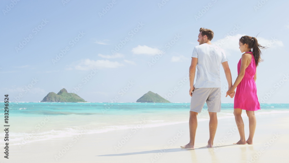 Wall mural Beach couple looking at ocean view from behind. Couple standing on white sand in pink dress and beachwear on vacations on Lanikai beach, Oahu, Hawaii, USA with Na Mokulua Islands. SLOW MOTION.