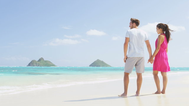 Beach couple looking at ocean view from behind. Couple standing on white sand in pink dress and beachwear on vacations on Lanikai beach, Oahu, Hawaii, USA with Na Mokulua Islands. SLOW MOTION.
