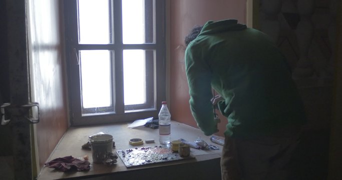 Man Artist in Green Clothes is Standing Close to Window at Windowsill is Preparing the Paints for Painting Indoors
