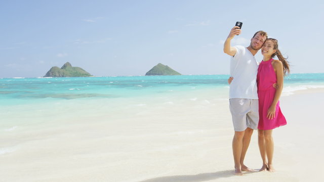 Couple on beach vacation taking selfie photograph using smart phone. Romantic couple in love on honeymoon on Lanikai beach, Oahu, Hawaii, USA with Mokulua Islands. Couple holding smartphone camera.