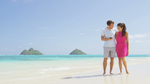 Couple on beach vacation using smart phone app. Romantic couple in love on honeymoon having fun on Lanikai beach, Oahu, Hawaii. Couple holding smartphone. RED EPIC SLOW MOTION.