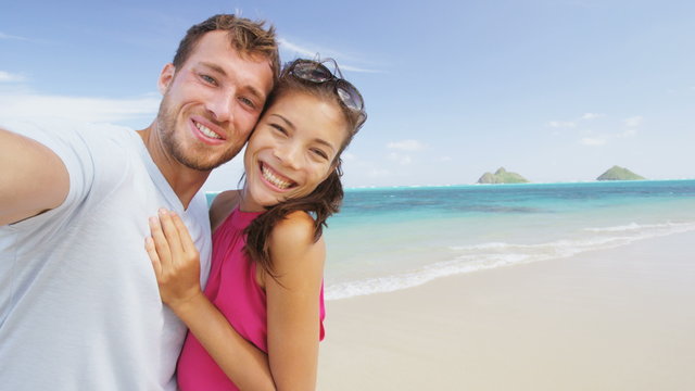 Couple on beach vacation taking selfie photograph using smart phone. Romantic couple in love on honeymoon on Lanikai beach, Oahu, Hawaii, USA with Mokulua Islands. RED EPIC SLOW MOTION.