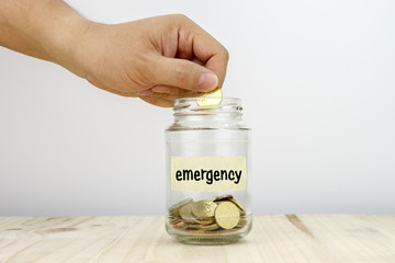 Hand putting a coin into a glass jar with word EMERGENCY on it.