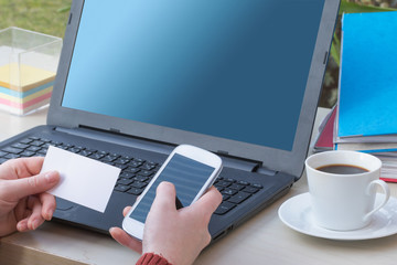 Closeup view of the woman`s holding a blank business card and smart phone in her hands. Notebook, folders and white cup of coffee are on the office table.