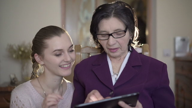 Mature woman in glasses sitting, using the tablet, girl coming to her in a room