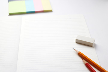 Blank notebook with pencil, red pencil, tag papers,  and eraser on white background.
