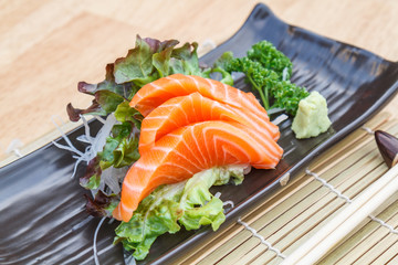 Sashimi salmon with vegetable and radish in black plate and chopsticks on wooden table.