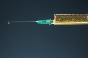 Hypodermic needle with a drop at its tip and syringe with dark yellow drug, on dark blue background