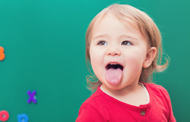Happy toddler girl sticking her tongue out in front of a chalkboard © Tierney