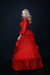 full length portrait of a beautiful blonde woman wearing a historical red silk, victorian era ball gown.