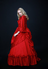 full length portrait of a beautiful blonde woman wearing a historical red silk, victorian era ball gown.