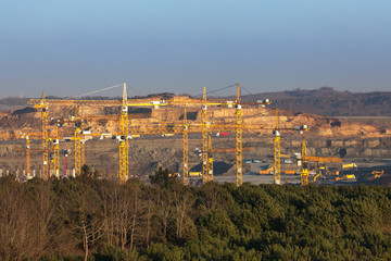 Cranes and trucks in a very big construction site near the forest.Many trees has been cut .