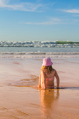 Kind sitzt im Sand und schaut entspannt den Wellen des Meeres zu, wie diese an den Strand rollen.