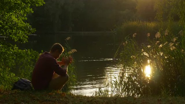 Man is Sitting at The Lake Bank Hands Are Folded Head Inclined Down Man is Praying Backpack is Close to Man Rippling Water Sun Reflection in Water