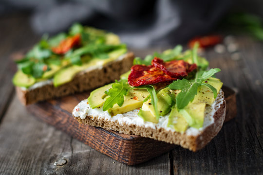 Healthy toast with goats cheese, avocado, arugula and sun dried tomatoes on rustic wooden table. Selective focus