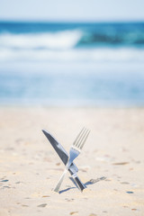 Fototapeta na wymiar Crossed processed knife and fork stuck in sand, shallow depth of field.