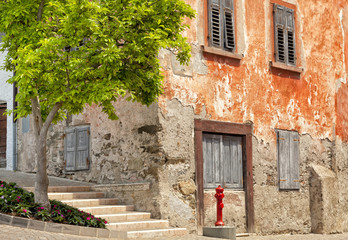 Old red historic corner house with aged walls and shutter windows, on cobbled street, with red water hydrant and tree in front