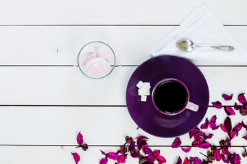 cup of tea or coffee on violet plate, silver spoon, marshmallow in glass vase, lump sugar, violet purple dry decor scattered on white colored wooden table,  top view