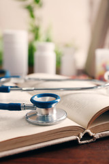 Book, bottles of pills and stethoscope on wooden table closeup