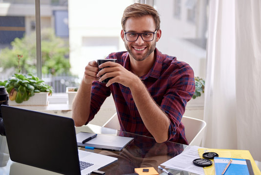 Young professional holding coffee and smiling at his desk