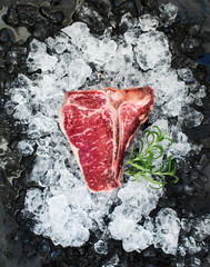 Raw fresh meat t-bone steak on chipped ice with rosemary over dark slate stone backdrop