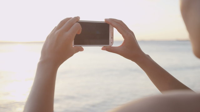 Tourist taking photograph of sunset on beach. View from woman using smartphone. Mobile cell smart phone girl outside.