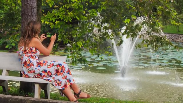 Blond Girl Sits on Bench at Fountain Takes Photos with Iphone