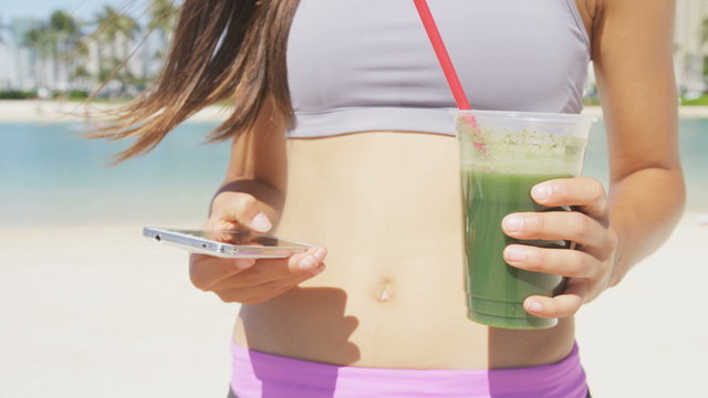 Smart phone fitness woman drinking green vegetable smoothie juice using smartphone app after workout running training. Fit female girl living healthy wellness lifestyle on summer beach.