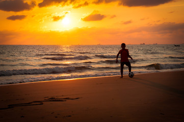 Silhouette of young boy playing a football on the beach in sunse
