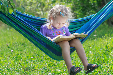 Concentrated two years old girl reading opened book on hanging hammock in green summer garden...