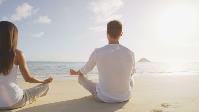 Yoga people meditating in lotus pose relaxing outside on beach at sunrise. Couple woman and man in meditation in serene ocean landscape. Lanikai beach, Oahu, Hawaii, USA.