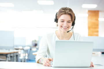 Happy young woman sitting and working with laptop using headset