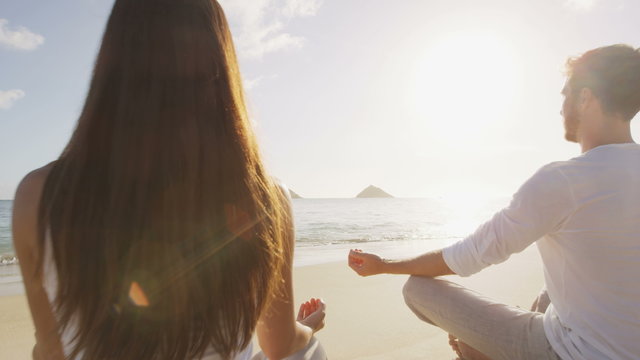 Yoga people meditating in lotus pose relaxing outside on beach at sunrise. Couple woman and man in meditation in serene ocean landscape. 