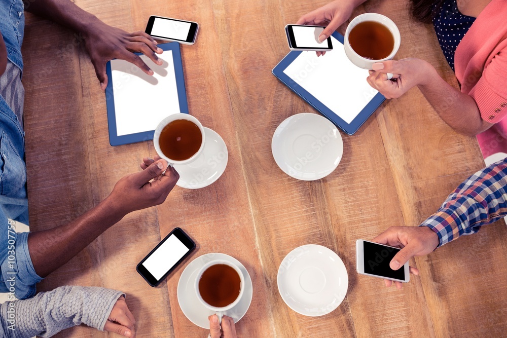 Poster colleagues using technologies at desk while holding coffee cups