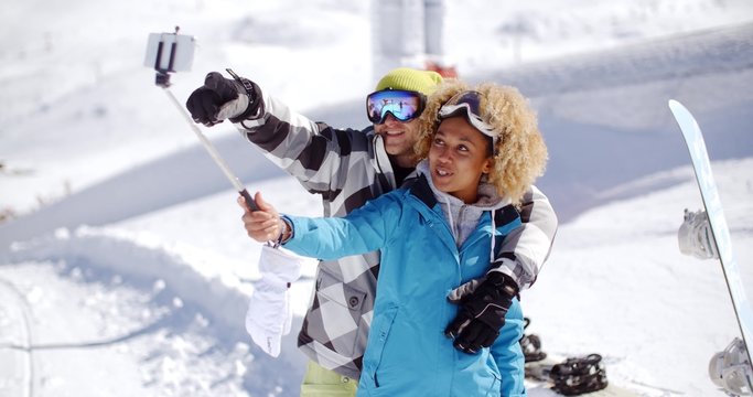 Fun young couple wearing ski clothes and goggles posing in the snow for a selfie on their mobile phone using a stick at a winter resort with their snowboard.