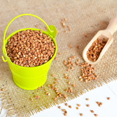 A small green bucket with raw buckwheat on a burlap and a white wooden background