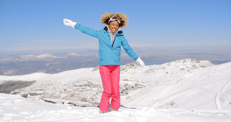 Happy beautiful young adult female in pink and blue skiing outfit waving arms around in the air near top of hill with mountain in background