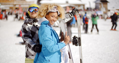 Fototapeta na wymiar Man in goggles flirting with smiling woman in blue jacket holding snowboard on crowded ski slope on sunny day