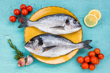 Fresh dorado fish on yellow plate and vegetables on blue background. Top view.