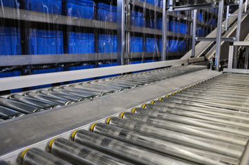 Close up shot of two roller conveyors in an automated warehouse with a ramp and piles of blue plastic boxes in the background. Photo taken in a big warehouse in Germany. Focus on the conveyor rolls.