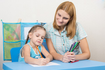 The five-year child draws on a sheet of paper, next to the teacher holds pencils