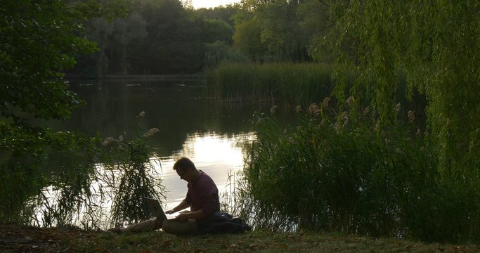 Man in Glasses Sits at The River Lake Holding the Laptop Working with Laptop Freelancer Programmer Copywriter Designer Accountant Stands Up Green Trees