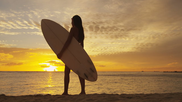 Surfer woman in silhouette walking with surfboard at sunset on tropical beach. Surfing girl looking at ocean sunset. Female bikini woman by water standing with surfboard in healthy active lifestyle.