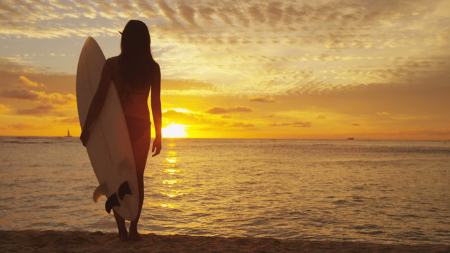 Young female surfer standing on beach sand under sunset sky holding surfboard watching ocean waves sunrise. Healthy lifestyle and sport concept. RED EPIC footage. 