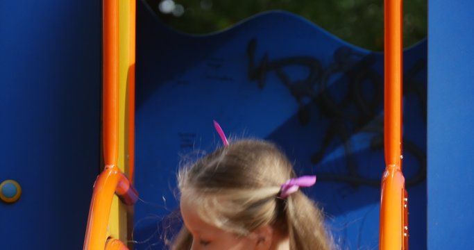 Girl is Sitting on The Chute With Pink Toy Tilda Rabbit Girl is Going Down Little Girl With Long Fair Hairs in Orange T-Shirt is Playing Playground