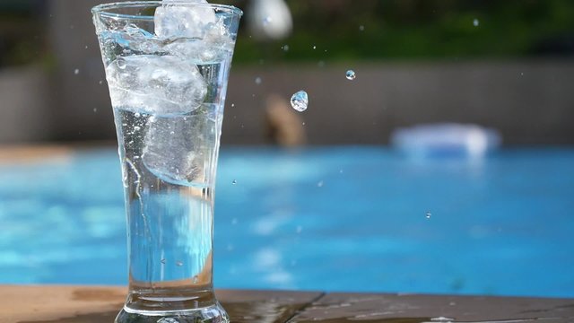 Ice Cubes Falling in Glass of Water near Swimming Pool. Slow Motion.