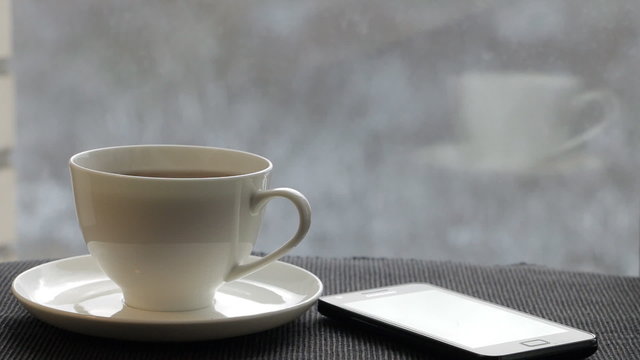 White  cup with steaming tea is in front of the window. Smartphone is nearby the cup. Winter and snowfall are outside.