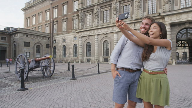 Stockholm tourists couple taking selfie picture with smartphone of Stockholm Royal Palace, Sweden, Europe. Happy tourist people sightseeing landmark attractions in Gamla Stan, the old town.
